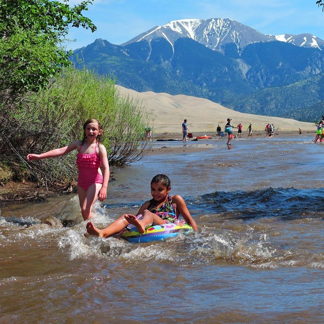Two girls float and run on a creek with a wave, with green trees on the sides of the creek