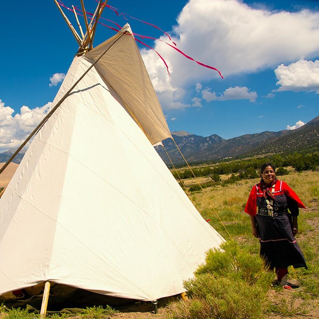 A Jicarilla Apache woman stands beside a tipi with the dunes and mountains in the background