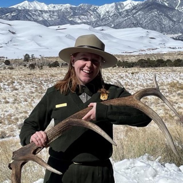 Ranger holding elk antler with snow covered mountains and dunes in the background