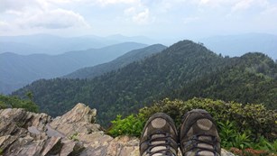 A hiker rests her feet at an overlook with the view of mountains extending to the horizon.