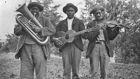 Three African American men are standing holding instruments - a tuba, guitar, and fiddle with a bow
