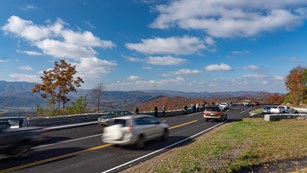 A roadway with pullouts to enjoy a mountain view. Cars blurred by motion move along the road.