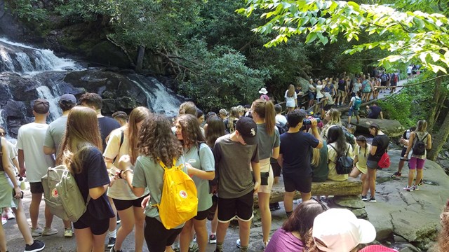 A large crowd of people gathered along a trail and on rocks near a waterfall surrounded by greenery.