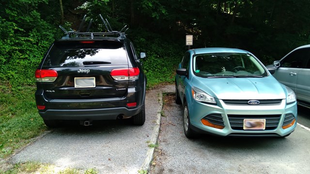   A black SUV parked on a gravel sidewalk next to a blue SUV parked on pavement in front of trees