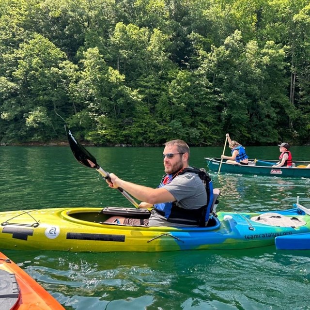 A kayaker on an adaptive kayak in a bright blue lake. Behind them is a canoe with three people. 