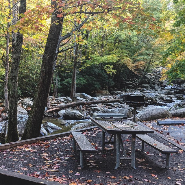 A wooden picnic table beside a river with large rocks and trees lining the sides