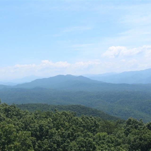 A partly cloudy sky above rolling blue-green mountains and dark green forests