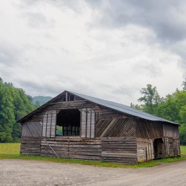 A wooden barn in a grassy field below a cloudy sky alongside a gravel road