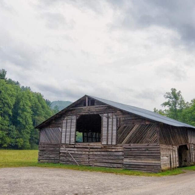 A wooden barn in a grassy field below a cloudy sky alongside a gravel road