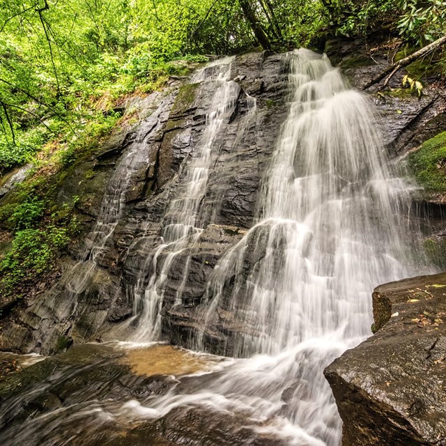 A waterfall cascading down rocks, some with moss and greenery. Green trees surround the falls.