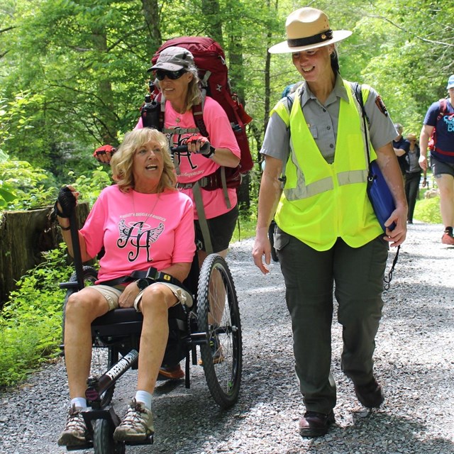 A person in a wheelchair on a trail beside a ranger.