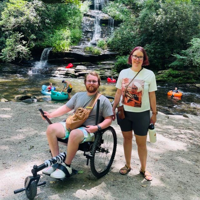 Two people pose for a picture on a trail in front of a waterfall 1 standing, 1 in a wheelchair