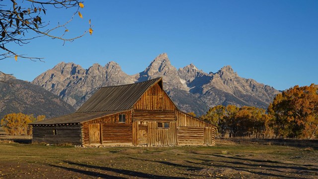 A brown, wooden barn sits in a valley in front of a mountain range.
