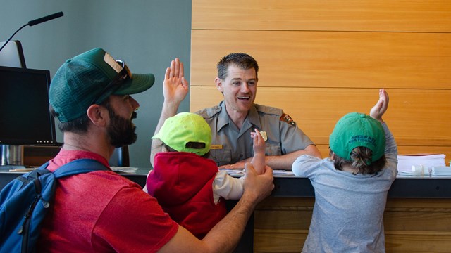Two kids are sworn in as Junior Rangers by a park ranger at a visitor center front desk.
