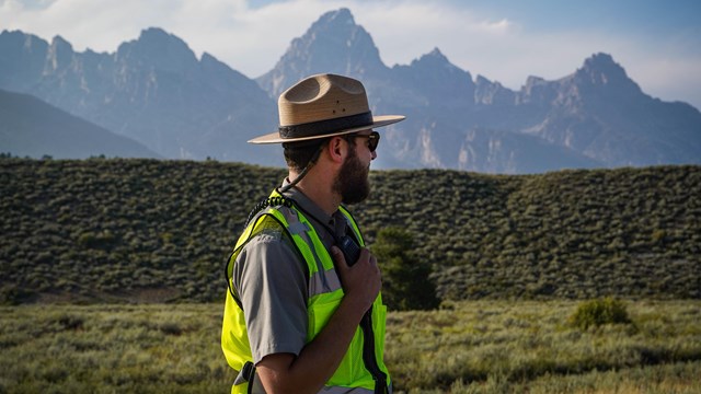 A park ranger in a safety vest communicates over the park radio with mountains in the background.