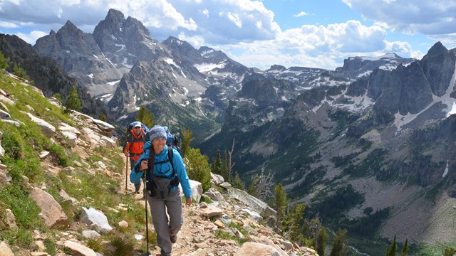 Two backpackers on a rocky trail in the Teton Range with the Grand Teton in the background.