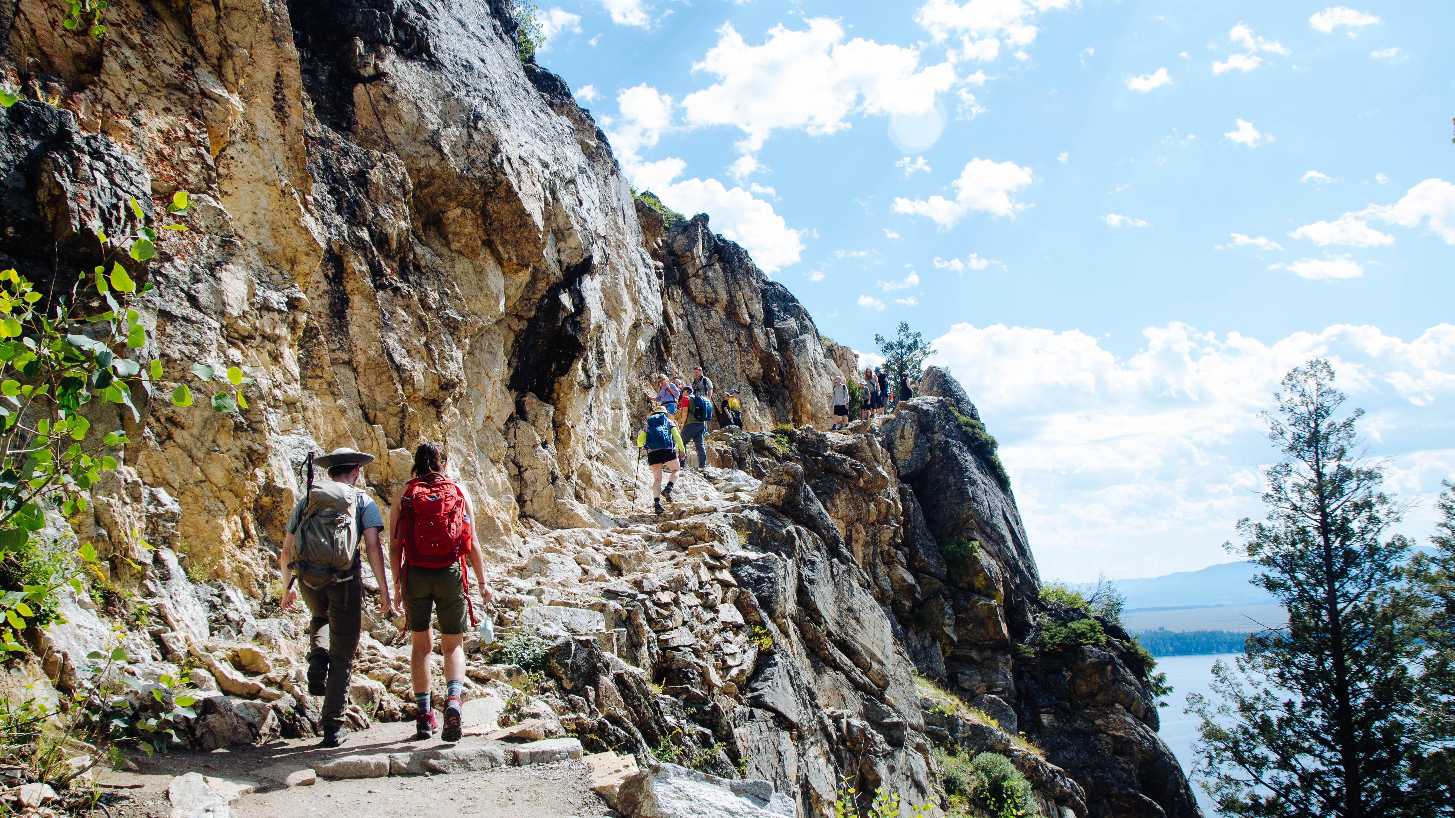 Two hikers walk up a rocky trail by a cliff.