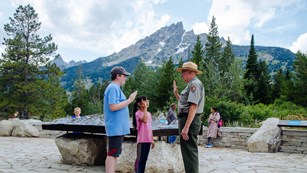A ranger stands with two kids with a mountain in the background.