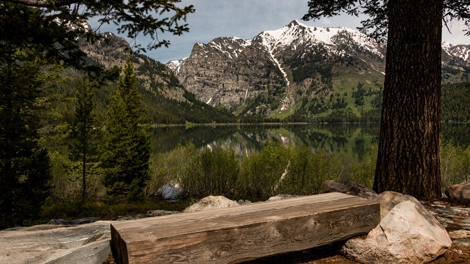 A wooden bench sits beside a lake surrounded by mountains.