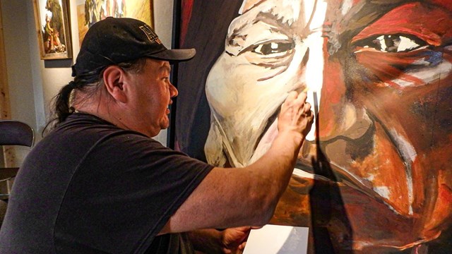 A seated man painting a large face on a canvas