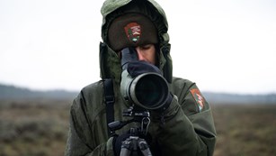 a ranger in a fleece hat looks through a spotting scope
