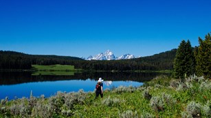 A hiker walks along a lakeshore with mountains in the background.