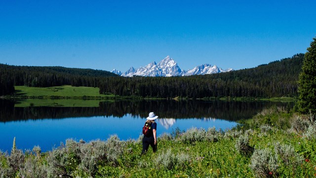 A hiker walks along a lake with mountains in the background.