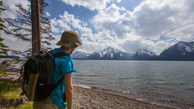 A young hiker looks across a large lake to view the Tetons on the other side