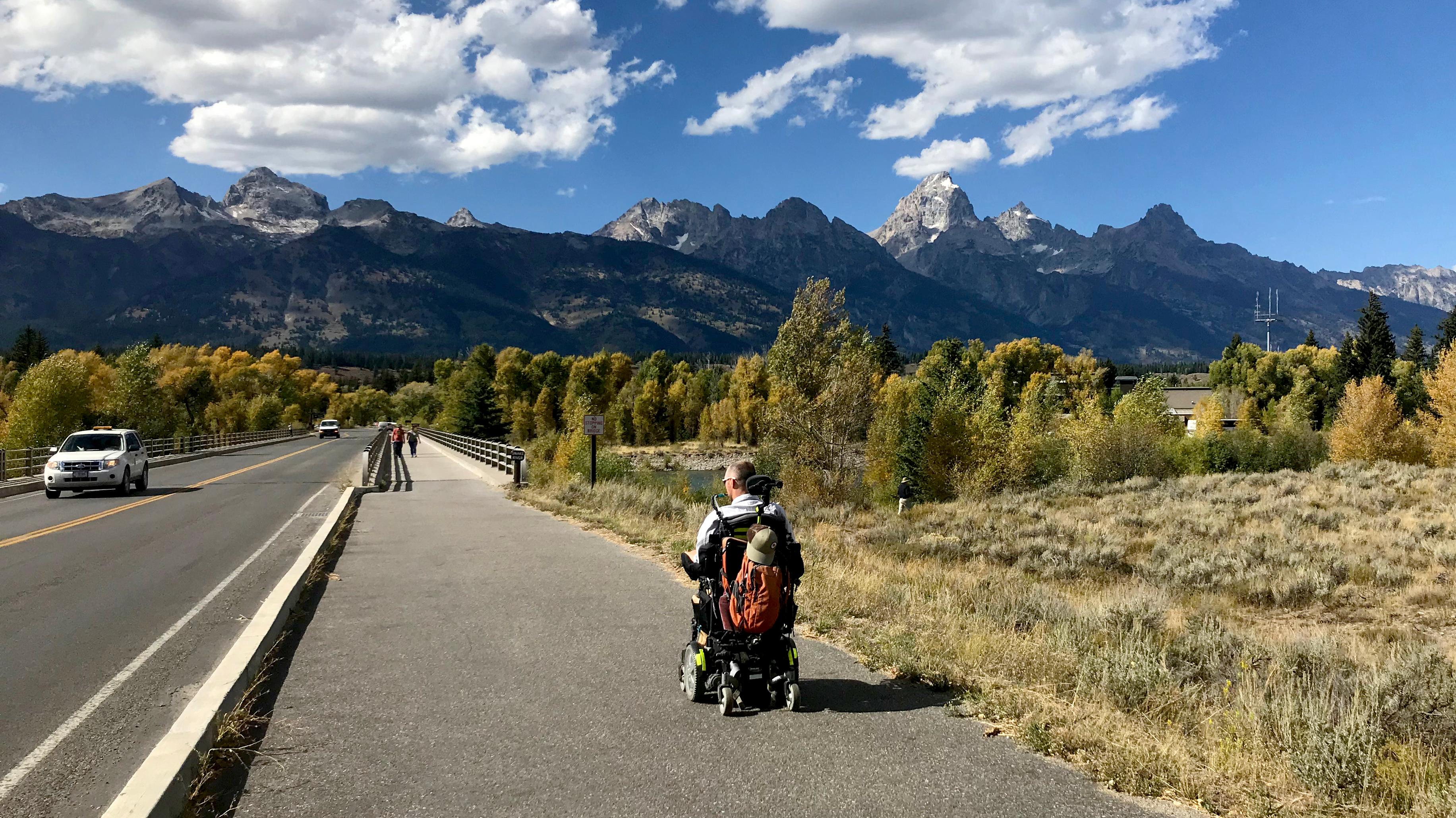 A visitor in a wheelchair on a paved path with mountains in the background.