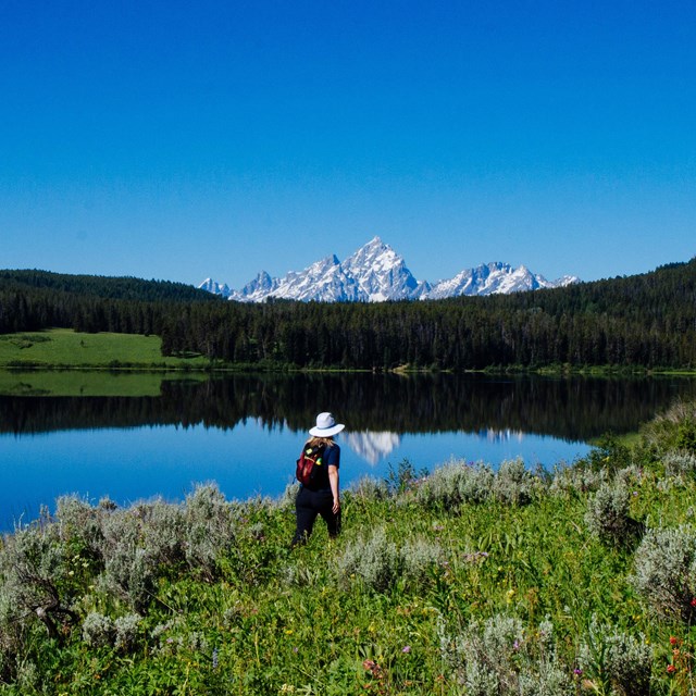 A hiker walks along a lake with mountains in the background.
