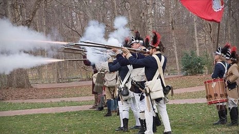 Infantry soldiers fire black powder muskets, boy with drum and boy with flag stand to rear.