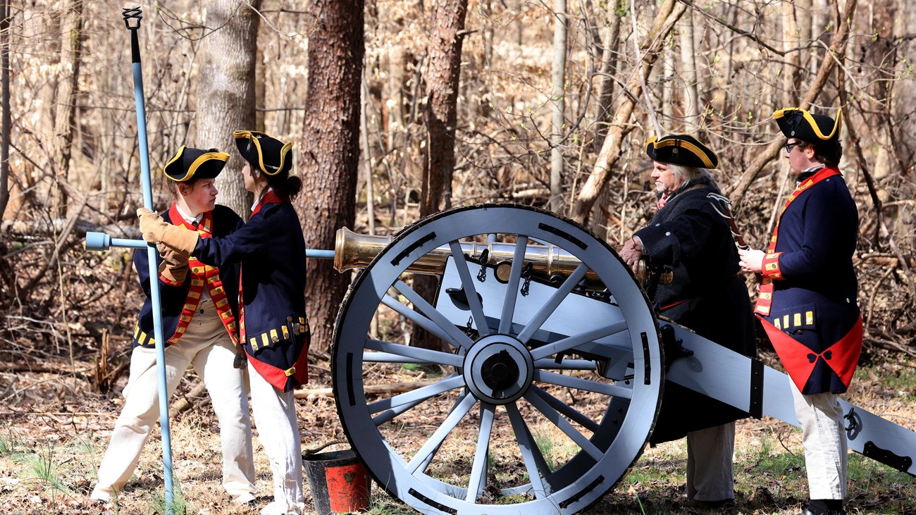 A bronze six pound cannon is being loaded. Four gunners in blue coats surround the piece.