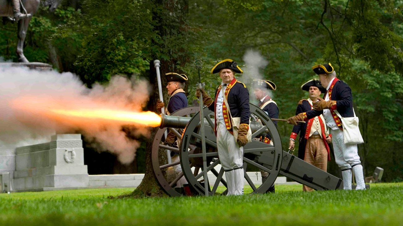 A bronze six pound cannon is firing a jet of flames. Five gunners in blue coats surround the piece.