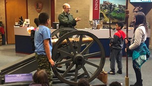 Park ranger teaches three students how to load a 3-pounder cannon and other students look at them.