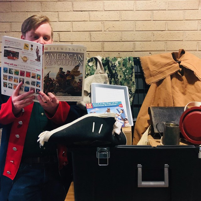 Man reads book about American Revolution, black trunk filled with soldier equipment