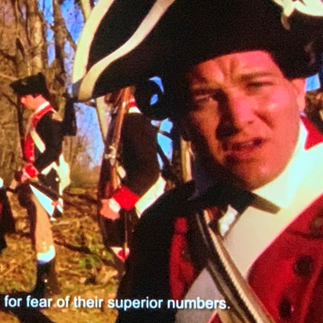 Continental soldier speaks to the camera while other continentals stand in camp. Wearing blue & red