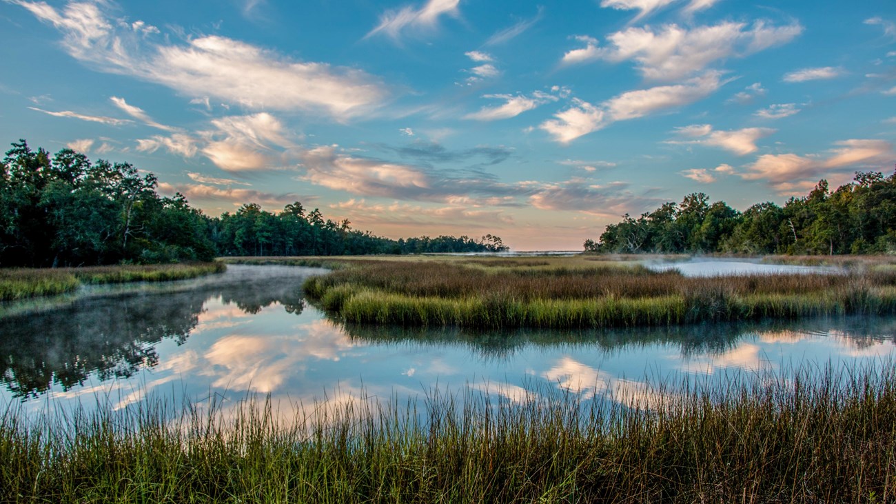 Clouds reflect over a still bayou with mist rising off the water at sunrise.
