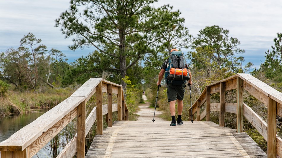 A man backpacking the Florida Trail hikes across a wooden footbridge onto the gravel trail beyond.
