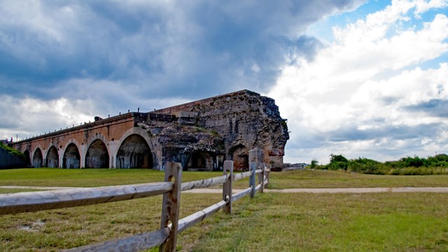 The corner of a fort stands on a grassy field with wooden fencing and clouds in the background.