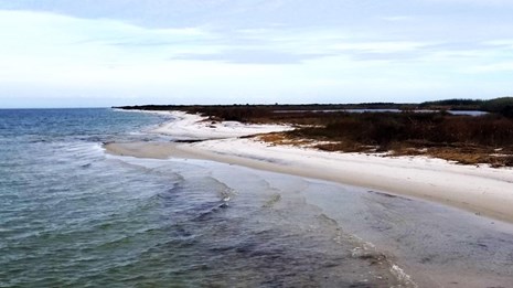 Small waves crash from left to right on to a barrier island with vegetated dunes to the right.