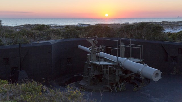 The sunsets over the water, in the foreground a cannon is mounted.