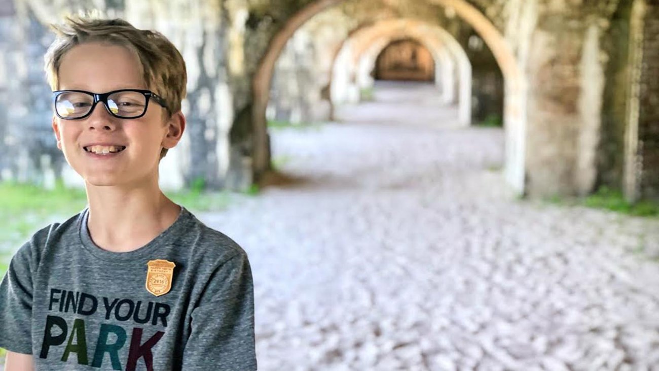 A child stands under a brick archways wearing a junior ranger badge.