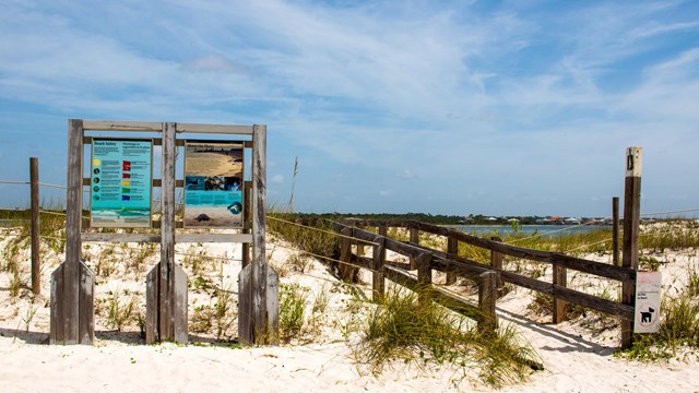 An informative sign and beach boardwalk are framed by a cloudy blue sky.