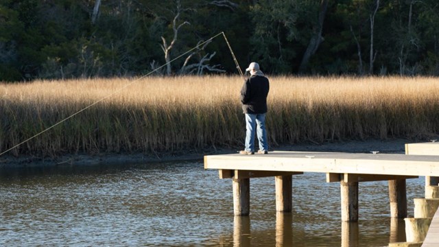 A fisherman casts a line into the bayou from a pier.