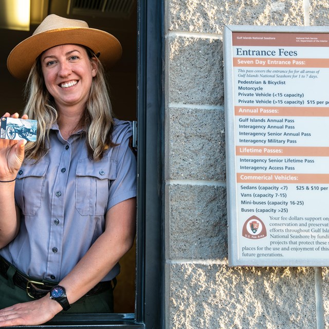 A ranger holding up an annual pass stands in the window of an entrance station.