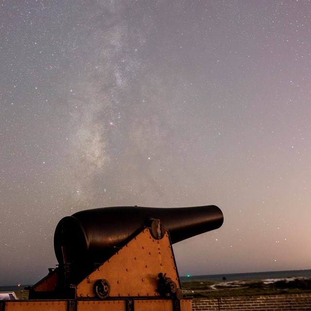 Stars can be seen in a night time photo, a large cannon is in the foreground illuminated.