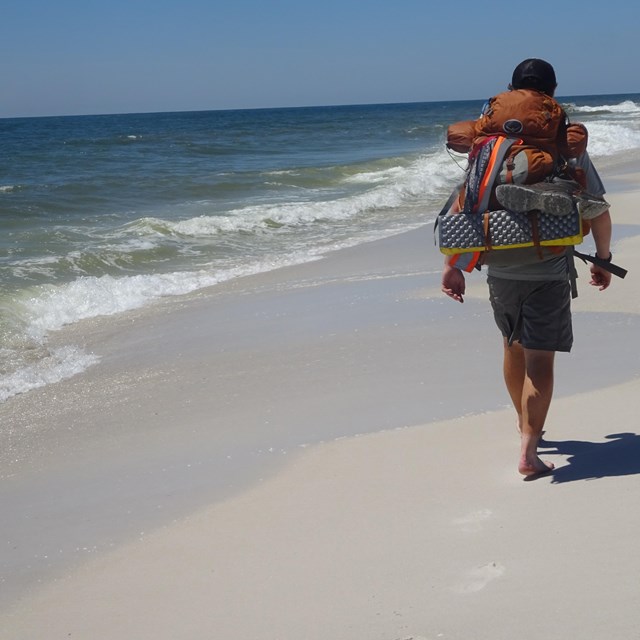 A lone backpacker walks along a sandy beach with water to his left.