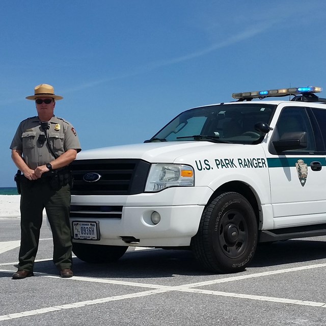 A park ranger stands in front of a patrol SUV just in front of a beach.