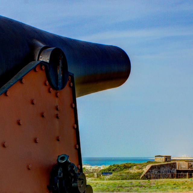 A large cannon sits on top of a historic fort with the coast in the background.