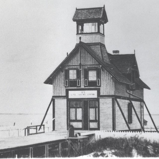 A black and white image of a narrow, two-story building on the waters edge.